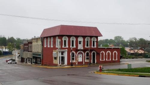 Historic red brick building with a red roof at a street corner, surrounded by small shops on a rainy day.