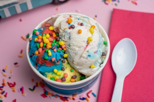 A colorful ice cream cup with three scoops topped with vibrant sprinkles, set against a pink background.