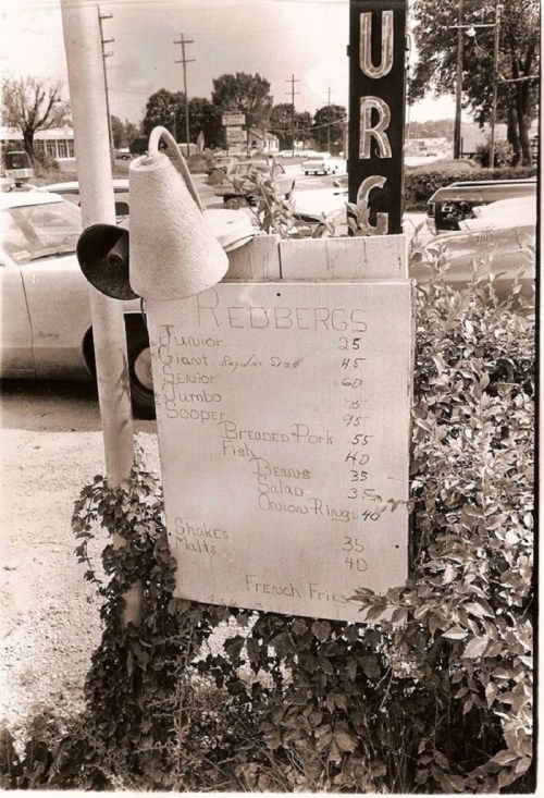 A vintage menu board listing food items and prices, with a hanging bell and cars in the background.