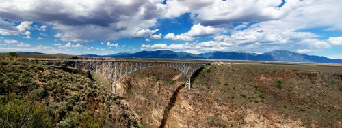 A panoramic view of a bridge spanning a deep canyon, surrounded by mountains and a blue sky with fluffy clouds.