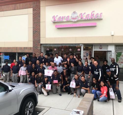 A large group of people, including police officers and community members, gather outside Kara Kakes bakery, smiling and holding treats.