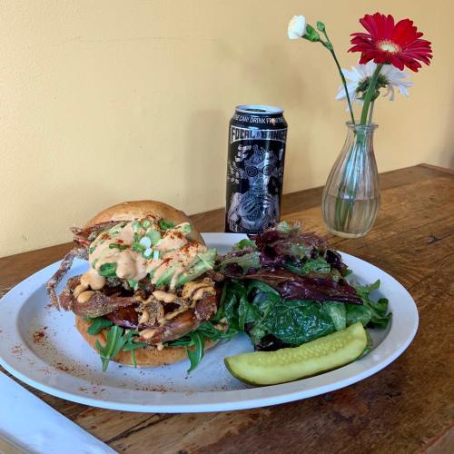 A hearty sandwich with greens, sauce, and pickles, served with a side salad and a can of Focal Banger beer.