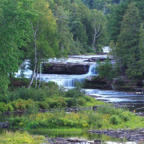 A serene landscape featuring a flowing river with waterfalls, surrounded by lush green trees and rocky formations.