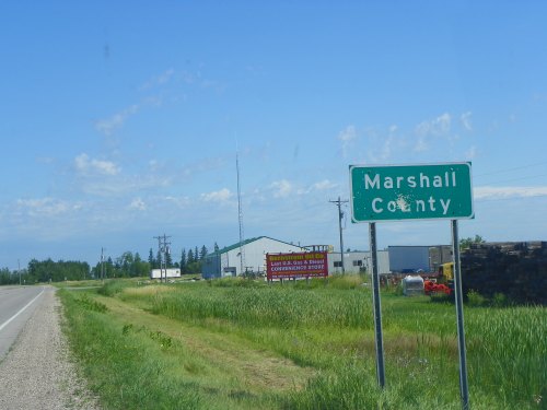 Sign for Marshall County along a rural road, with green grass and buildings in the background under a clear blue sky.