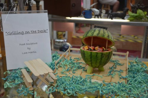 A watermelon carved as a grill, filled with colorful fruit, displayed on a table with a sign about the sculpture.