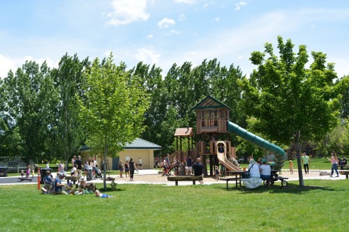 A sunny park scene with a playground, trees, and families enjoying the outdoors on a grassy area.