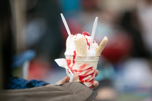 A hand holding a cup of ice cream topped with bananas, whipped cream, and cherry sauce, with two spoons.