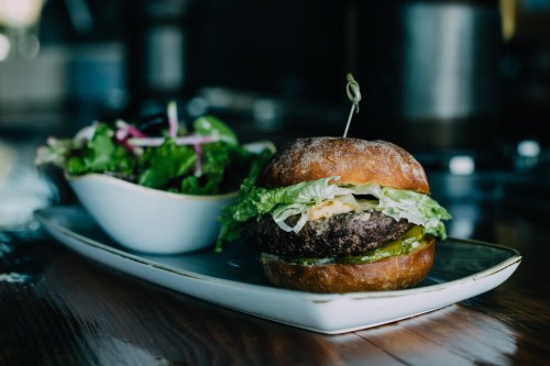 A juicy burger with lettuce and pickles on a plate, accompanied by a side salad in a white bowl.