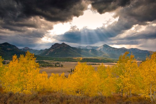 A dramatic landscape featuring golden autumn trees, mountains, and dark storm clouds with rays of sunlight breaking through.