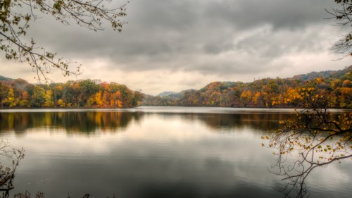 A serene lake surrounded by autumn foliage under a cloudy sky, reflecting the colorful trees in the water.