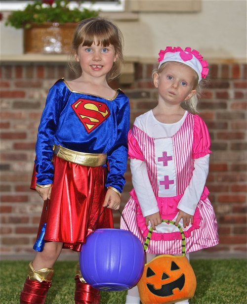 Two young girls in Halloween costumes: one as a superhero and the other as a nurse, holding candy buckets.