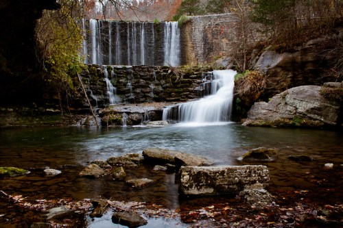 A serene waterfall cascades over rocks into a calm pool, surrounded by autumn foliage and stone walls.