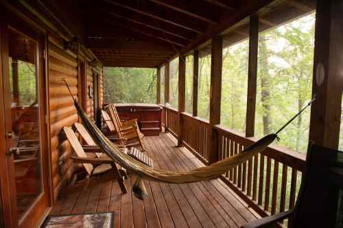 A cozy wooden porch with a hammock and several chairs, surrounded by lush greenery.