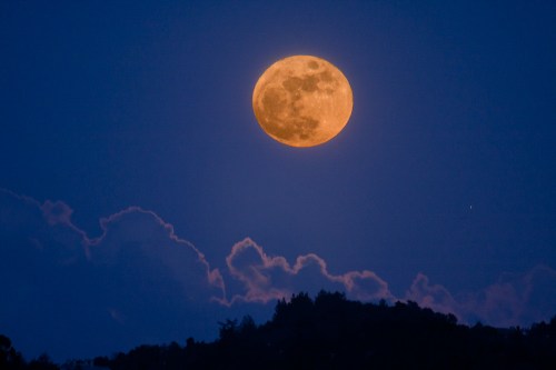 A large, glowing full moon rises above a dark silhouette of hills and clouds against a deep blue sky.