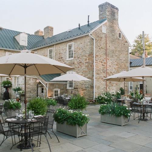 A charming outdoor dining area with stone buildings, umbrellas, and lush greenery in planters.