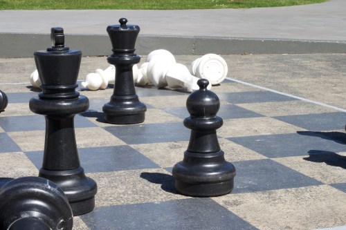 A close-up of a chessboard with large black pieces and fallen white pieces on a sunny day.