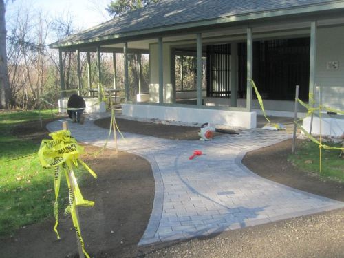 A partially completed pathway with yellow caution tape near a building, surrounded by grass and trees.