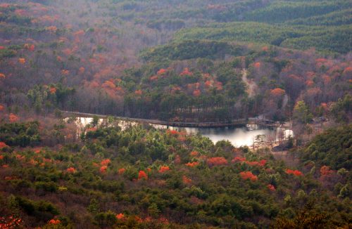 Aerial view of a forested landscape with a winding lake, surrounded by autumn foliage in shades of red and green.