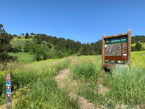 A grassy trail leads to a sign for Hansen-Larsen Memorial Park, surrounded by trees and hills under a clear blue sky.