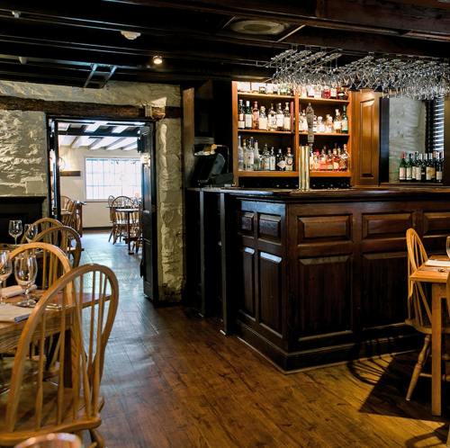 Cozy restaurant interior featuring a wooden bar, shelves of drinks, and wooden tables with chairs. Warm lighting enhances ambiance.