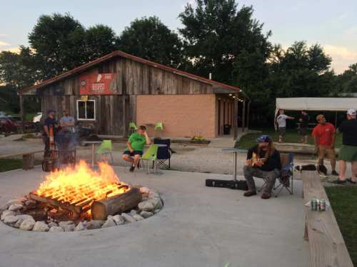 A cozy outdoor gathering with a campfire, people relaxing, and a musician playing guitar near a rustic building.