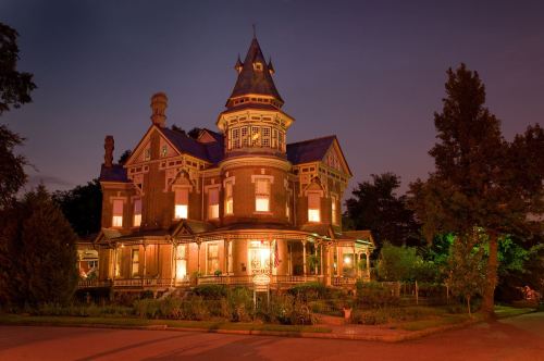 A beautifully lit Victorian-style mansion at dusk, featuring intricate architecture and lush greenery.