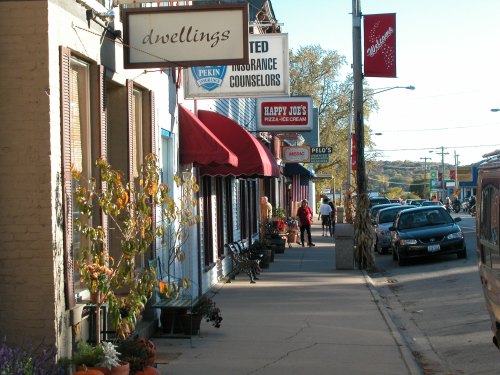 A quaint street scene featuring shops with awnings, parked cars, and pedestrians enjoying a sunny day.