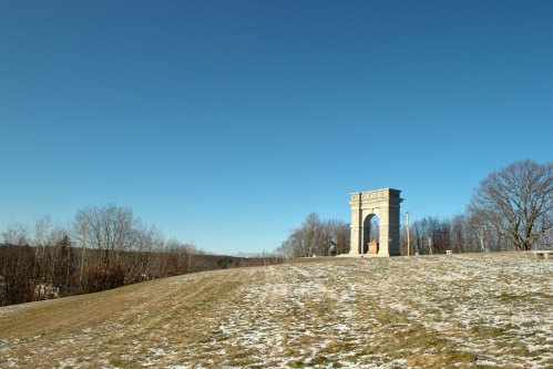 A stone archway stands on a snowy hillside under a clear blue sky, surrounded by bare trees.