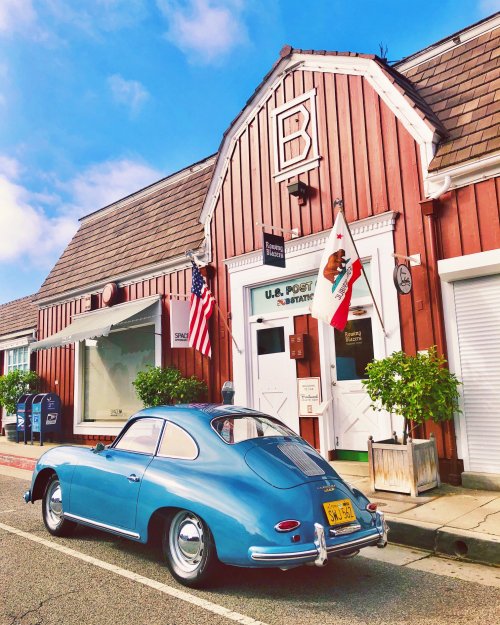 A vintage blue car parked in front of a red building with flags and a post office sign.