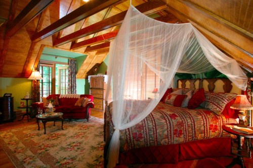 Cozy attic bedroom with a canopy bed, floral decor, and warm lighting, featuring a sitting area and wooden beams.
