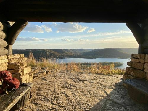 View from a rustic shelter overlooking a lake and rolling hills at sunset, with a backpack resting on a bench.