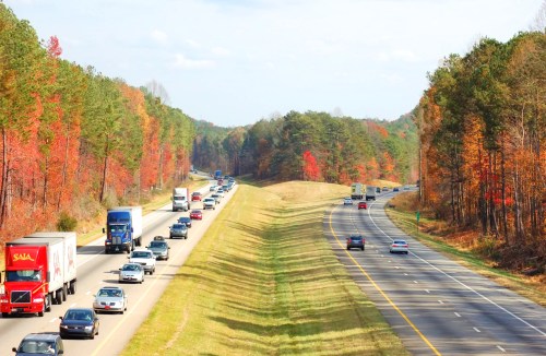 Traffic flows on a highway surrounded by trees displaying autumn colors.