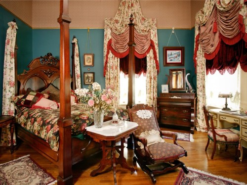 A vintage bedroom with floral decor, a wooden bed, a rocking chair, and a dresser, featuring elegant curtains and a cozy atmosphere.