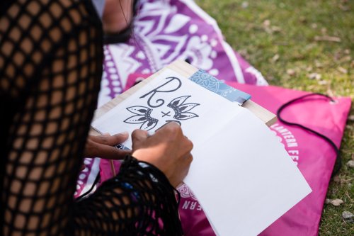 A person sketches a floral design with the letter "R" on a blank page, sitting on a colorful mat outdoors.