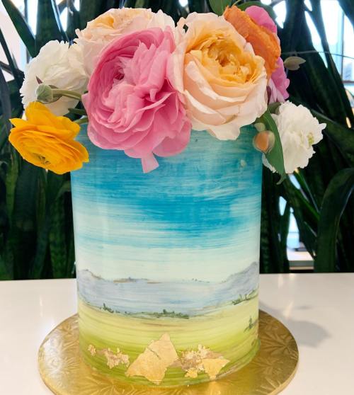A beautifully decorated cake with a painted landscape and topped with colorful flowers, set against a green backdrop.