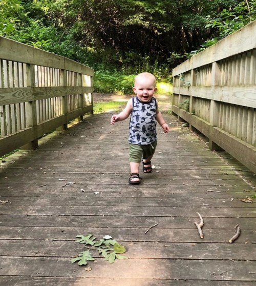 A toddler walks confidently on a wooden bridge surrounded by greenery, smiling and enjoying the outdoors.