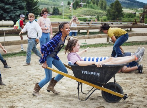 A girl pushes a wheelbarrow with a laughing friend inside, while other kids play in a sandy outdoor setting.