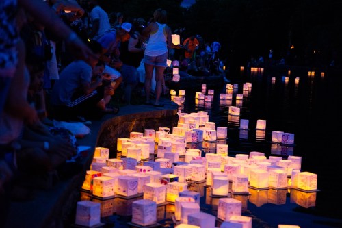People gather by a pond at dusk, releasing glowing lanterns that float on the water's surface.
