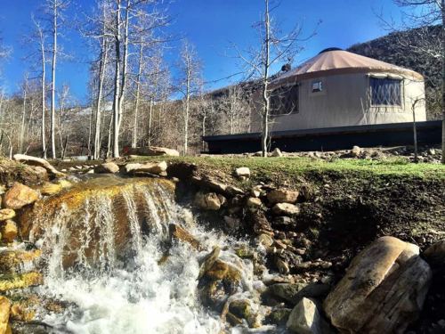 A yurt beside a flowing stream, surrounded by trees and mountains under a clear blue sky.