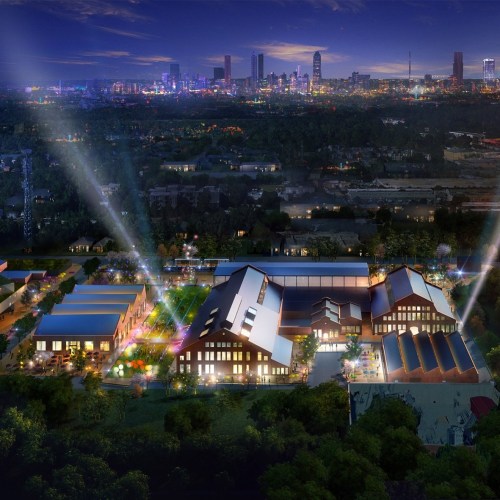 Aerial view of a modern complex at night, illuminated with lights, set against a city skyline in the background.
