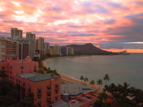 A vibrant sunset over a beach with palm trees, city buildings, and Diamond Head in the background.