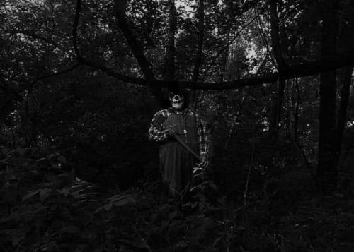 A shadowy figure in overalls and a skull mask stands in a dark, wooded area, holding a weapon.