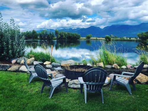 A serene lakeside scene with four chairs around a fire pit, surrounded by greenery and mountains under a cloudy sky.