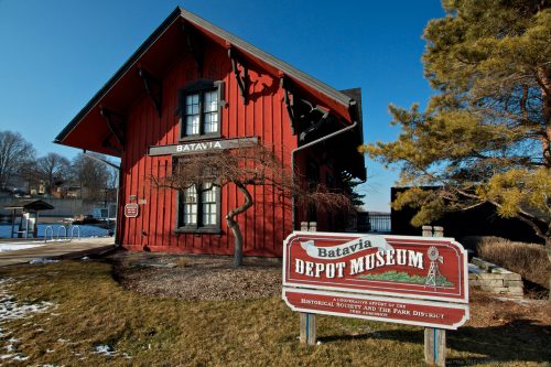 Red wooden building with a sign reading "Batavia Depot Museum," surrounded by grass and trees on a clear day.