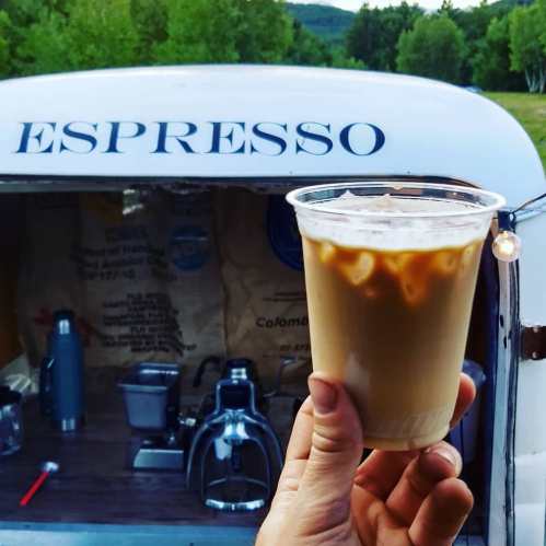 A hand holds a cup of iced espresso in front of a vintage espresso trailer with "ESPRESSO" written on it.