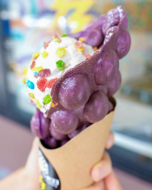 A hand holding a purple bubble waffle cone filled with purple ice cream and topped with whipped cream and colorful sprinkles.