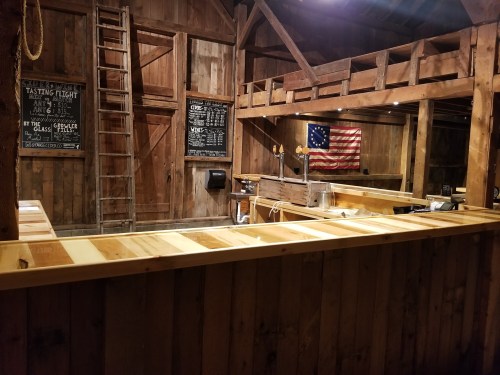 Interior of a rustic barn with wooden walls, a bar area, chalkboard menus, and an American flag hanging.