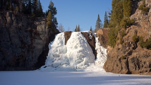 Frozen waterfall cascading over rocky cliffs, surrounded by evergreen trees and a snowy landscape under a clear blue sky.