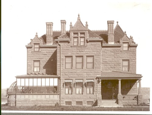 Historic stone mansion with a tiled roof, multiple chimneys, and a glass conservatory on the side.