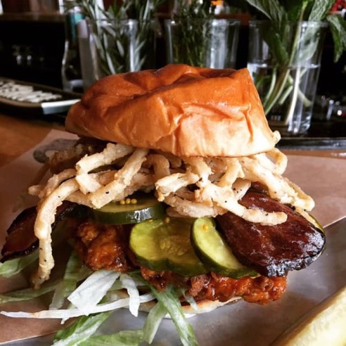 A hearty sandwich with fried chicken, pickles, lettuce, crispy onions, and a soft bun, served with a pickle on the side.